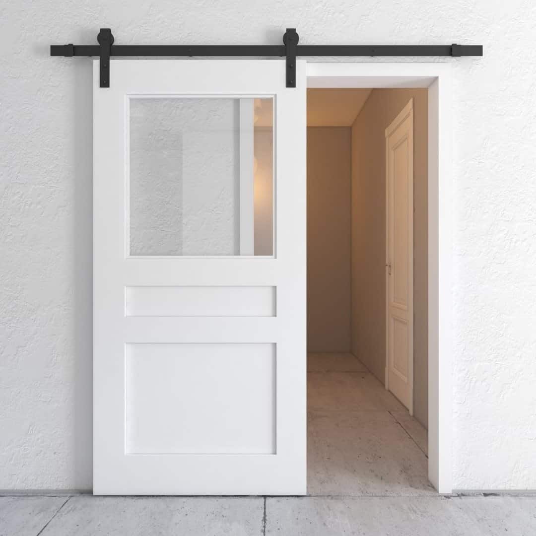 A white sliding barn door with glass panel and black hardware, partially open to reveal a hallway with white walls and closed doors.