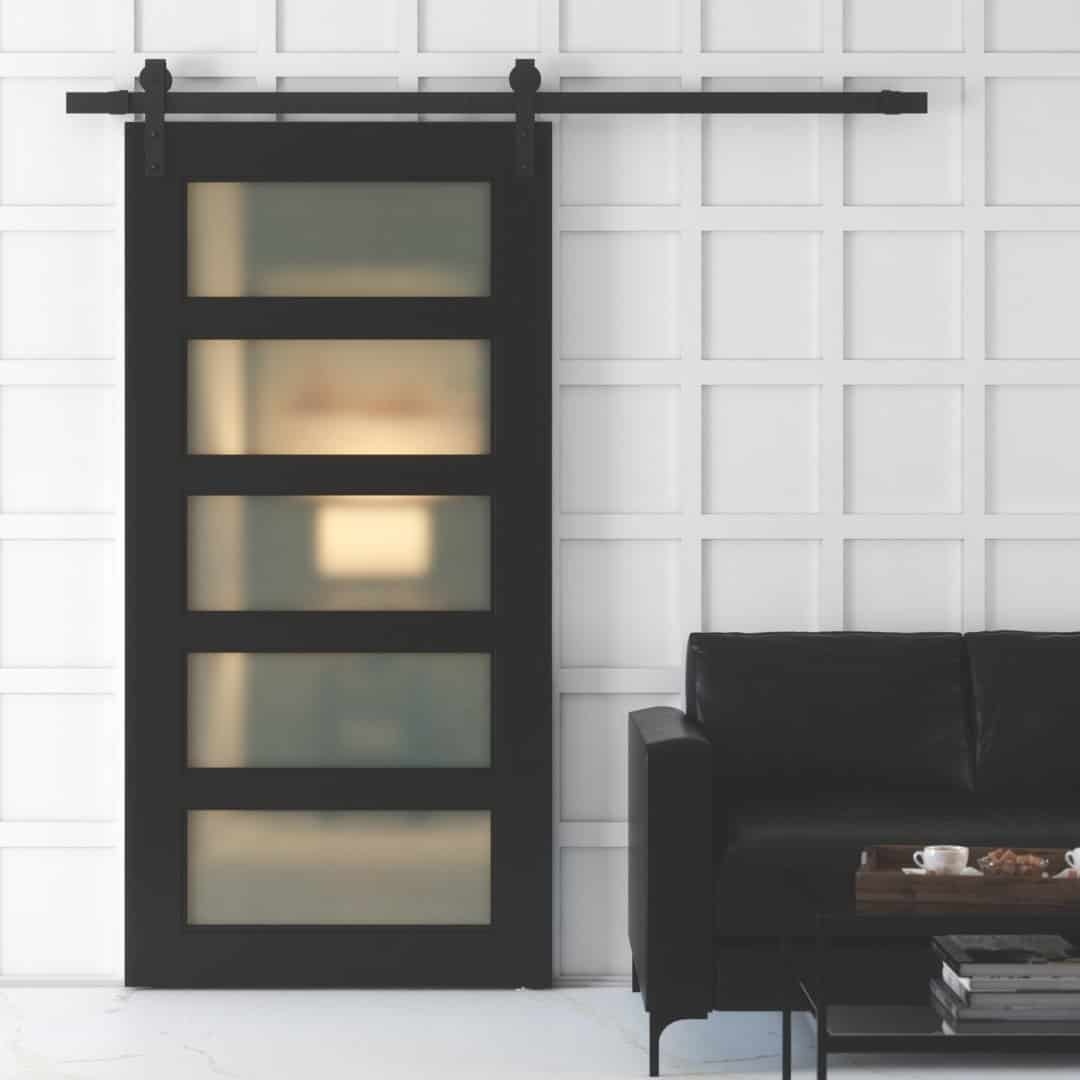 A Boston 5 Panel Barn Door with frosted glass panels, mounted on a white wall next to a black leather sofa with a coffee cup on the armrest.