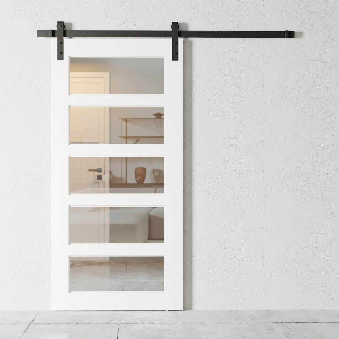 Urban Woodcraft Boston 5 Panel Barn Door - White Clear Glass (1) A white 5-panel sliding barn door with four glass panels, mounted on a black track, partially open to reveal a room with minimalistic decor.