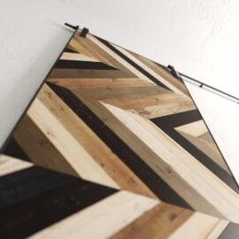 A Chevron 83" x 40" Barn Door showcasing an intricate geometric chevron pattern in multiple wood shades, mounted diagonally against a white wall.
