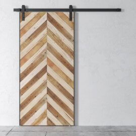 The Herringbone 83" x 40" Barn Door, featuring a chevron pattern made from wooden planks in varying shades, is mounted on a black metal track system above a concrete floor.