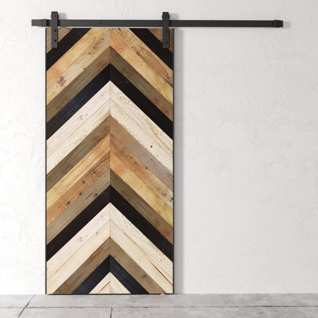 The Herringbone 83" x 40" Barn Door, featuring a striking chevron pattern with wooden panels in various shades of brown, tan, and black, is beautifully mounted on a white wall.