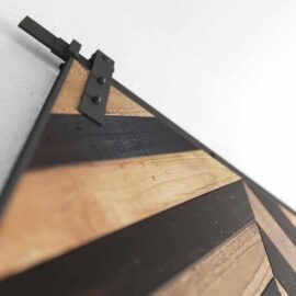 Close-up of the Herringbone 83" x 40" Barn Door, showcasing a diagonal pattern of alternating light and dark wood panels framed in metal. The metal slider mechanism is visible at the top of the door.