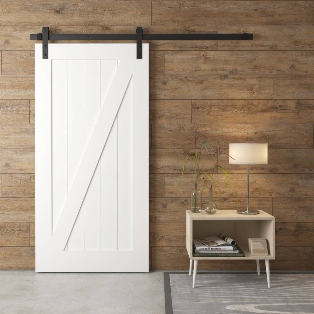 A Z-Panel Barn Door in white on a wooden wall beside a small table with a lamp, books, and a glass vase.