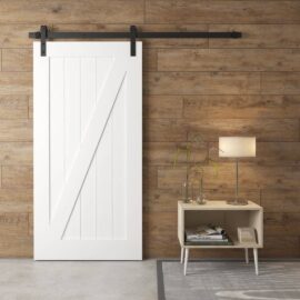 A Z-Panel Barn Door in white on a wooden wall beside a small table with a lamp, books, and a glass vase.