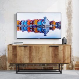 A flat-screen TV displaying colorful houses is mounted on a wall above the Parisian Factory 4 Door 72″ TV Console, which holds a stack of books and a small decorative object.