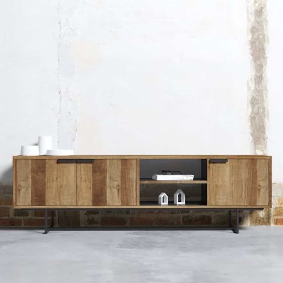The 3 Door Parisian Factory 78″ TV Console, with a charming rustic finish, boasts three cabinets and two open shelves perfect for displaying small decorative items. It stands elegantly against a partially plastered white wall with a brick base.