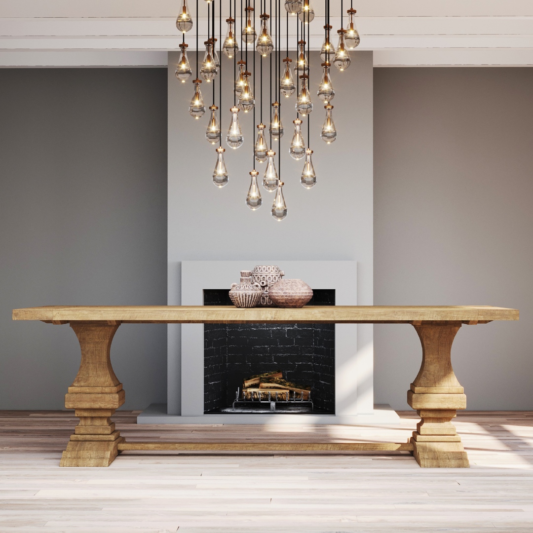 The Castello Dining Table - Natural, adorned with two decorative centerpieces, stands in front of a dark fireplace and white mantel, beneath a chandelier of hanging bulbs in a modern, minimalist room.