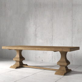 The Castello Dining Table - Natural features a wooden rectangular top with two ornate pedestal legs, set on a concrete floor against a gray wall.