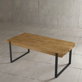 The Hudson Dining Table, featuring a rectangular wooden design with a jagged edge, sits on a concrete floor. It boasts black metal legs and a smooth top surface.