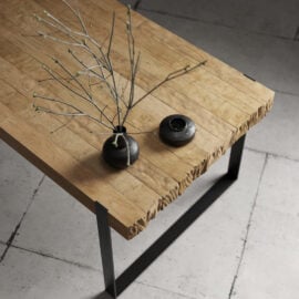 The Hudson Dining Table, a rectangular wooden piece featuring a raw edge, is beautifully adorned with a small branch in a black vase and complemented by a round black decorative item, all set atop a tiled floor.