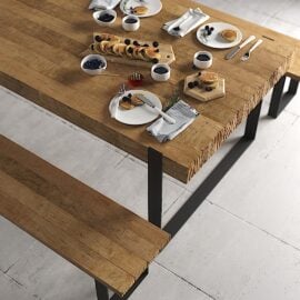 A Hudson Dining Table is set for breakfast with various small plates of food, bowls of berries, glasses, and cutlery. A cloth napkin is placed near the edge. The scene includes a wooden bench.