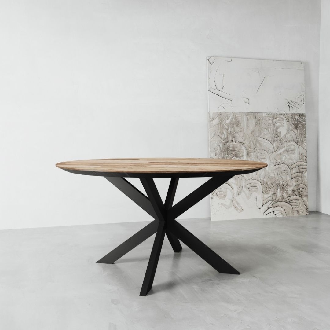 The Kolt Round Dining Table, featuring a wooden top and a black metal base, stands in a minimalist room with two large, partially visible abstract artworks propped against the wall.