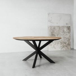 The Kolt Round Dining Table, featuring a wooden top and a black metal base, stands in a minimalist room with two large, partially visible abstract artworks propped against the wall.