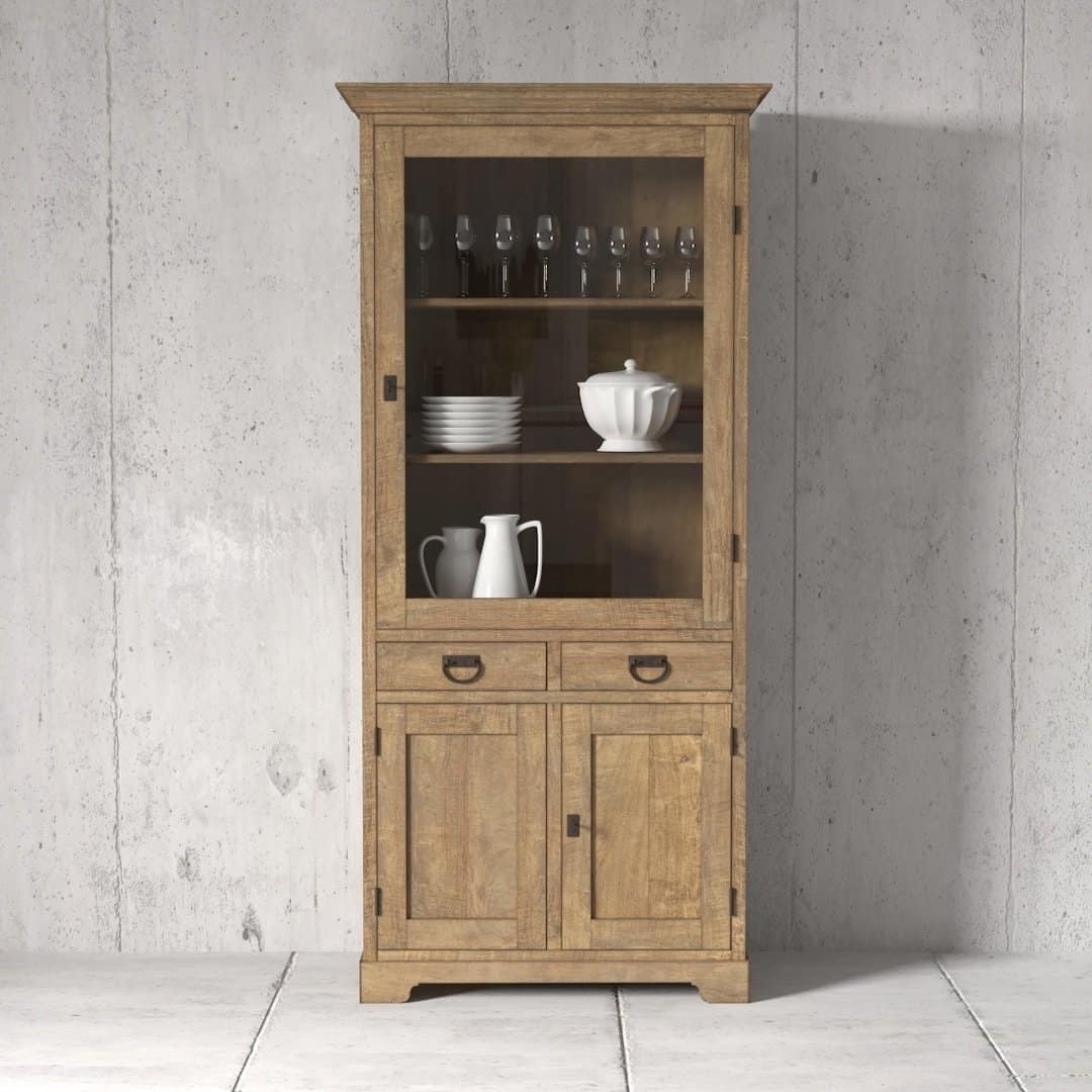 Riverbend Display Cabinet featuring a wooden frame with a glass door. This 36-inch unit showcases wine glasses, plates, and a teapot on shelves. Below, it offers two drawers and two closed cupboards, all set against a concrete wall.
