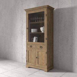 The Riverbend 36" Display Cabinet, made of wood and featuring glass doors, displays glassware and ceramic items on its shelves. It stands against a concrete wall and includes two drawers along with two closed compartments at the bottom.