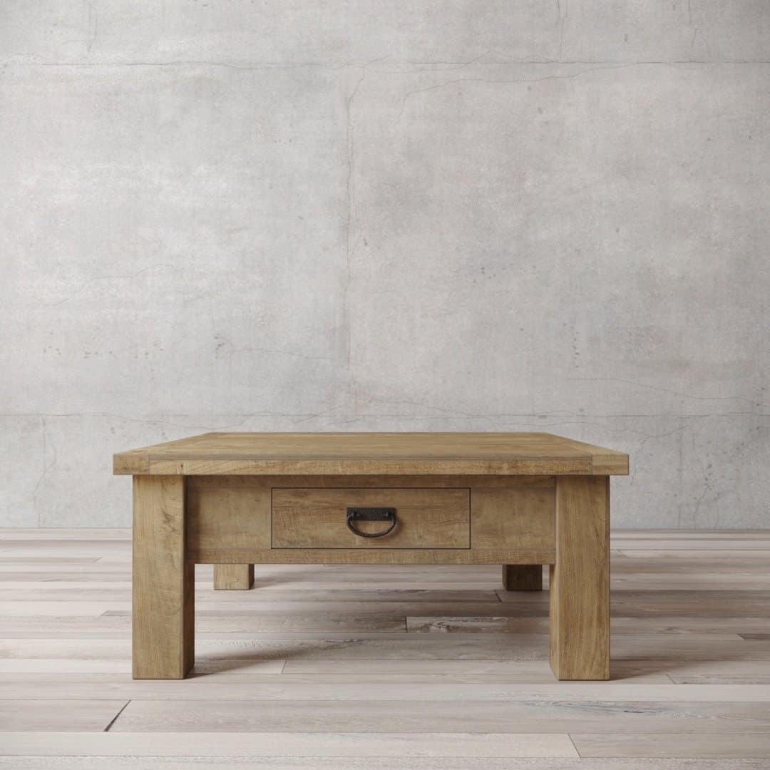 A Riverbend wooden coffee table with a single drawer is placed on a hardwood floor in front of a plain concrete wall.