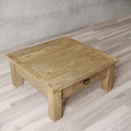 A Riverbend Coffee Table, square and wooden with a drawer, placed on a light-colored wooden floor against a plain concrete wall.