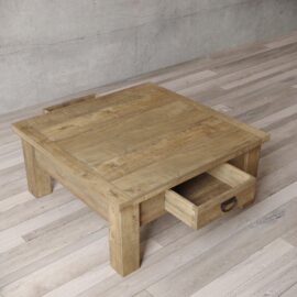 The Riverbend Coffee Table, featuring a wooden square design with thick legs and a single drawer, is showcased on a wooden floor against a plain wall.