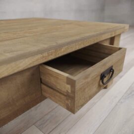 A close-up of the Riverbend Coffee Table with a drawer partially open, showcasing its dovetail joints and metal handle. The table features a natural wood finish with visible grain patterns.