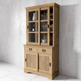 The Riverbend 36" Display Cabinet features a glass-paneled upper section showcasing glassware and a lower section equipped with drawers and sliding doors, all set against a plain concrete wall.
