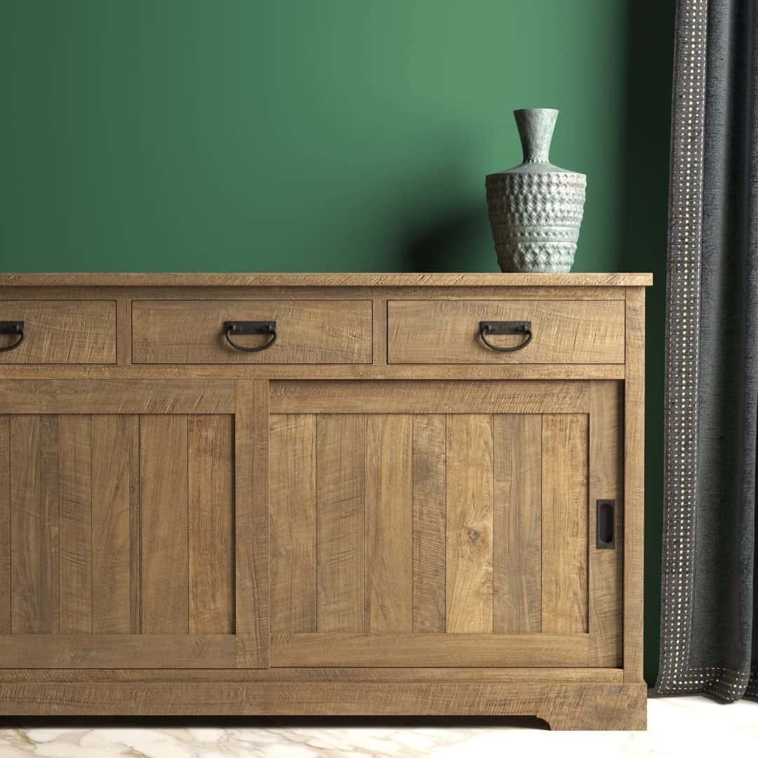 Urban Woodcraft 65 Riverbend Dining Buffet - Natural Livestyle The Riverbend Sliding Door Dining Buffet, featuring three drawers and a sliding door, stands against a green wall. A textured vase is placed on top, and a dark curtain is partially visible on the right.