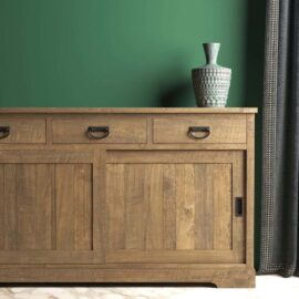 The Riverbend Sliding Door Dining Buffet, featuring three drawers and a sliding door, stands against a green wall. A textured vase is placed on top, and a dark curtain is partially visible on the right.