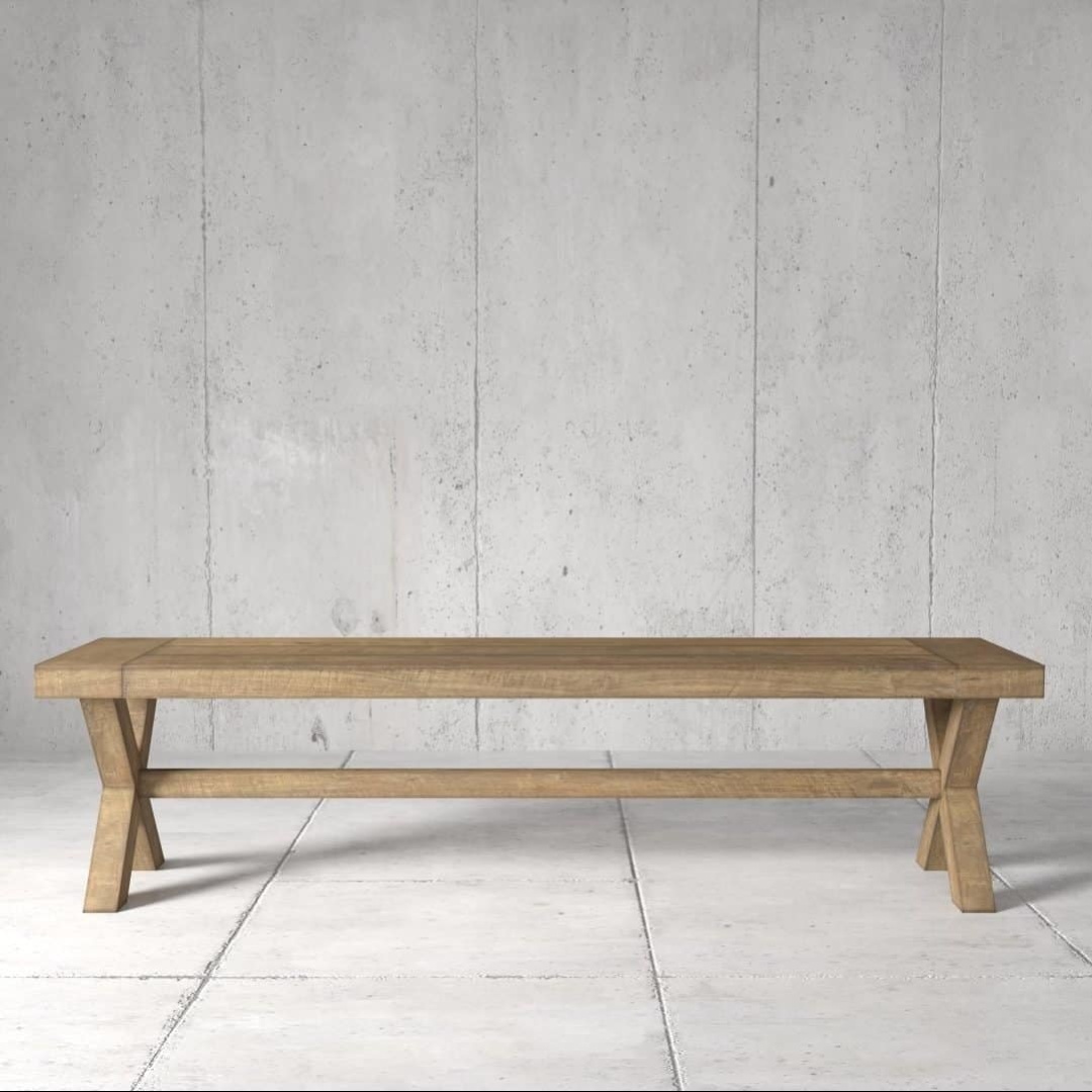 The Grand X Dining Bench, featuring an X-shaped leg design, sits on a concrete floor against a concrete wall.