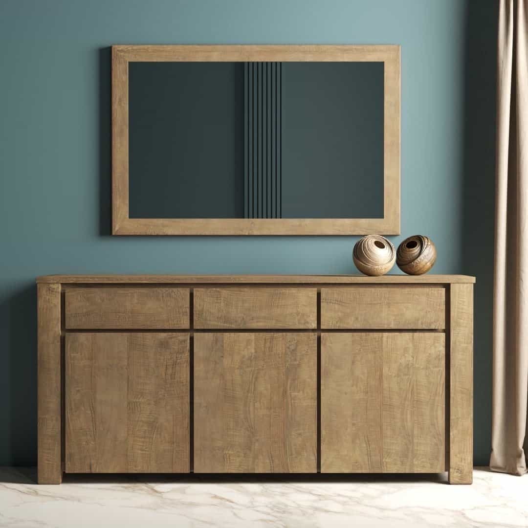 Urban Woodcraft 75 Teca Dining Buffet - Natural Lifestyle (1) The Teca Dining Buffet with three drawers and three cabinet doors stands against a teal wall, topped by a matching framed mirror and two decorative vases. A curtain is partially visible on the right.