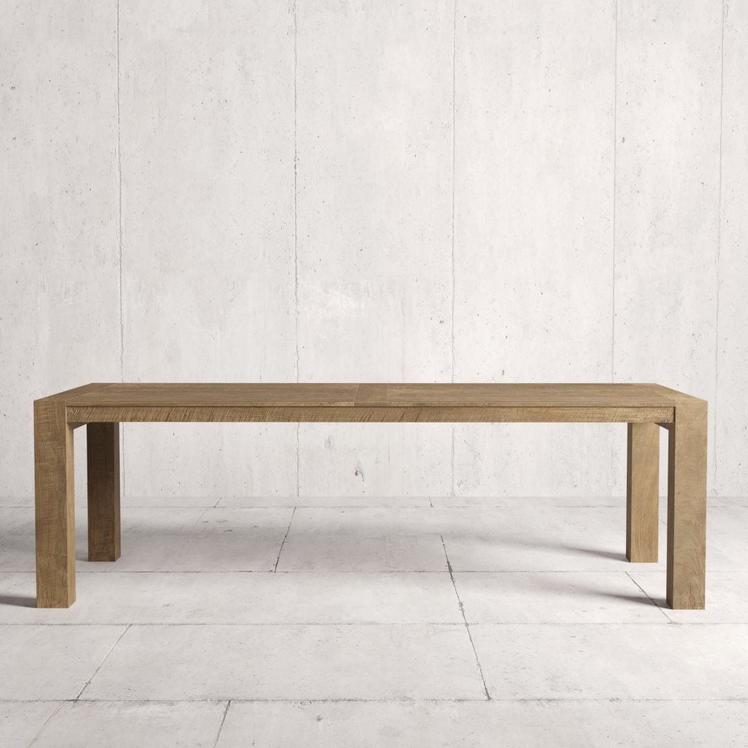 A long, rectangular wooden table with simple, clean lines, known as the Villa Dining Table, stands against a light-colored, textured wall and floor background. The natural finish highlights its elegance and timeless design.