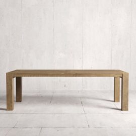 A long, rectangular wooden table with simple, clean lines, known as the Villa Dining Table, stands against a light-colored, textured wall and floor background. The natural finish highlights its elegance and timeless design.