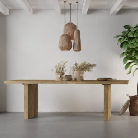 The Camden Dining Table adorned with decorative vases, pampas grass, and a bowl, sits under three hanging wicker lamps. A large green plant stands on the right against a white wall.