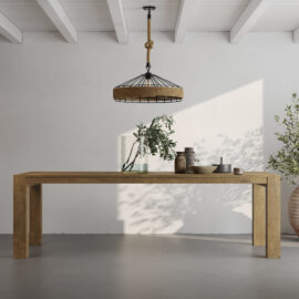 A minimalist room features the Villa Dining Table in natural wood adorned with decorative items, a hanging light fixture, and an olive tree plant against a white wall and concrete floor.
