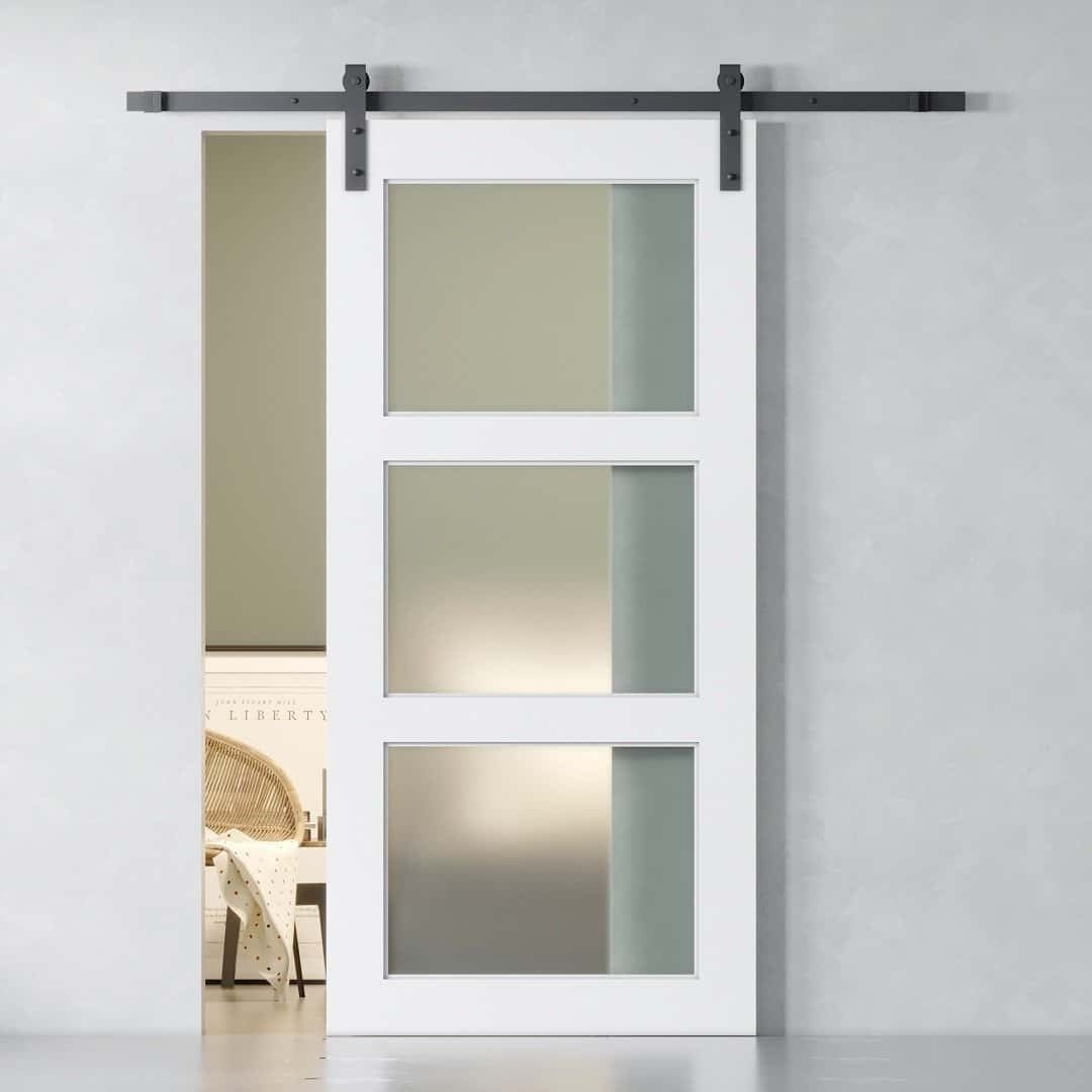 A white sliding barn door with 3-panel frosted glass partially reveals a room with a wicker chair and wall art. The door is elegantly installed on a metal rail system.
