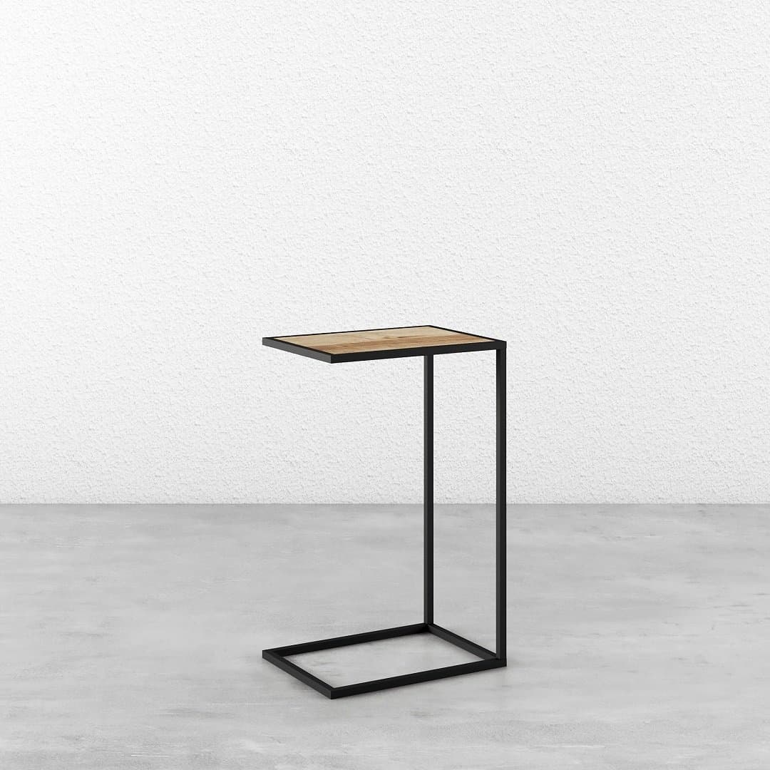 A Side Table with a black metal frame and light wood top, positioned against a white textured wall on a smooth grey floor.