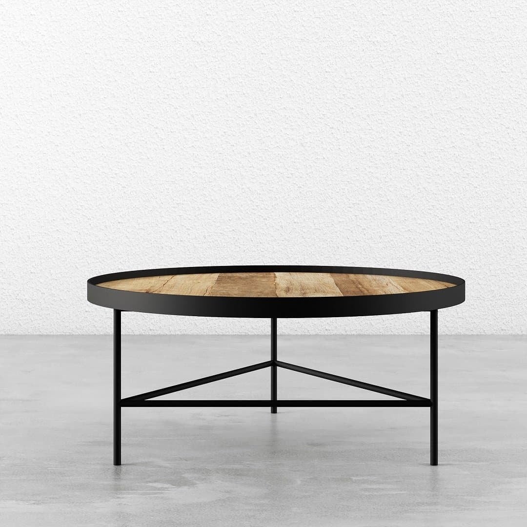 The Round Coffee Table, featuring a wooden top and black metal legs with a minimalist design, is placed on a concrete floor against a white textured wall.