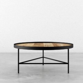 The Round Coffee Table, featuring a wooden top and black metal legs with a minimalist design, is placed on a concrete floor against a white textured wall.