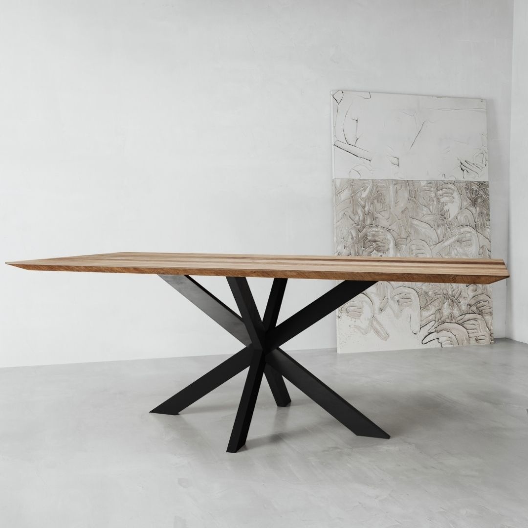 The Kolt Dining Table, featuring a sleek wooden top and a geometric black metal base, stands in a minimalist room with light gray walls and a concrete floor. Two large canvas artworks are leaned against the wall.
