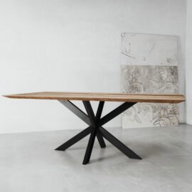 The Kolt Dining Table, featuring a sleek wooden top and a geometric black metal base, stands in a minimalist room with light gray walls and a concrete floor. Two large canvas artworks are leaned against the wall.