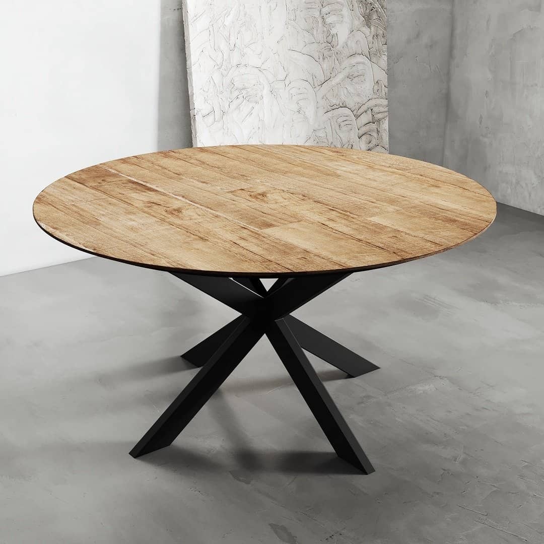 Round Kolt Dining Table 2 A round wooden dining table with a black metal X-shaped base is placed on a concrete floor against a light grey wall.