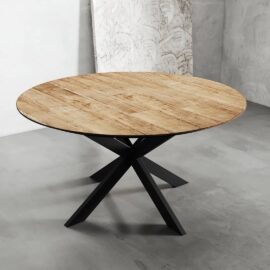 A round wooden dining table with a black metal X-shaped base is placed on a concrete floor against a light grey wall.