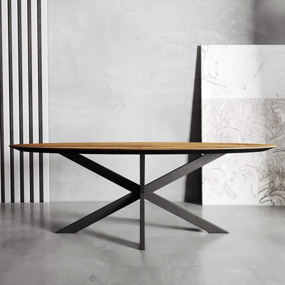 dtp 028 1 natural The Oval Kolt Dining Table, featuring a sleek oval wooden top and a black metal X-base, stands in a minimalist room with light gray walls and abstract wall art leaning in the background.