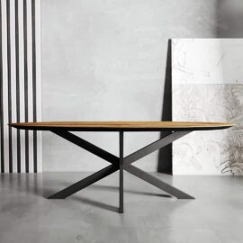 The Oval Kolt Dining Table, featuring a sleek oval wooden top and a black metal X-base, stands in a minimalist room with light gray walls and abstract wall art leaning in the background.