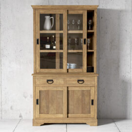 The Riverbend 45" Display Cabinet, featuring glass doors that elegantly showcase glassware, a pitcher, and bottles, stands against a concrete wall. The cabinet includes two drawers and two solid doors at the bottom for additional storage.