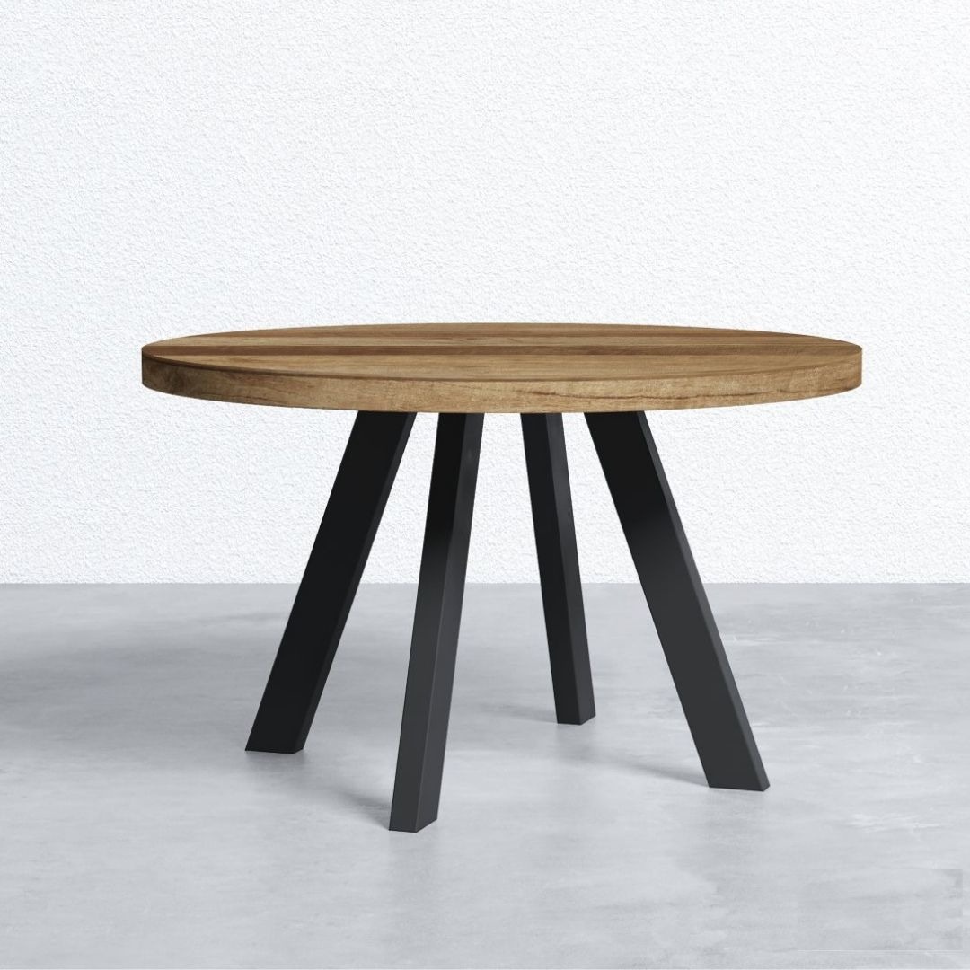 The Brickworks Round Table features a thick wooden top and four black metal angled legs, standing on a grey floor against a white wall.