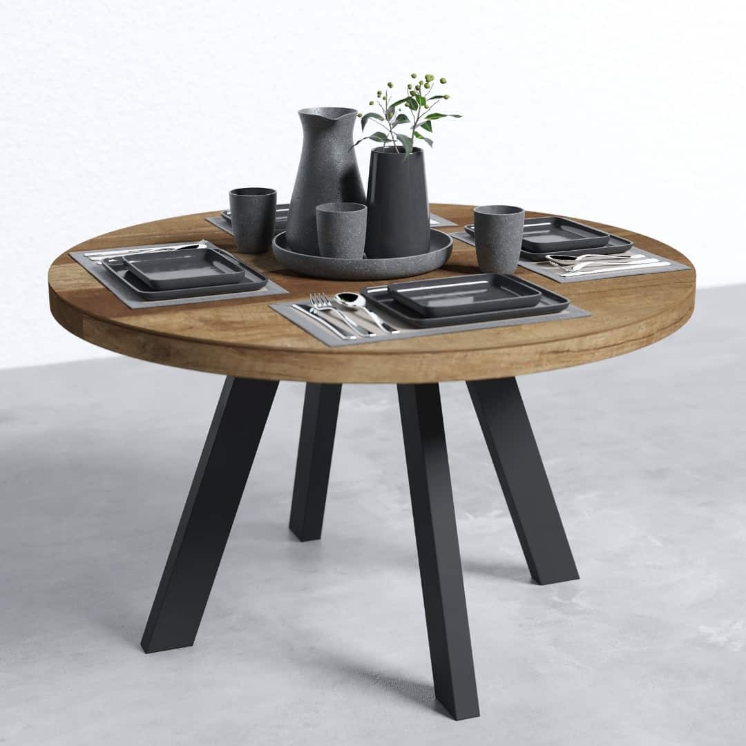 Natural Teak (1080 × 1080 px) The Brickworks Round Table, a round wooden piece with black metal legs designed for four people, is elegantly set with black stoneware dishes and cups, a matching pitcher, utensils, and a small vase adorned with greenery.