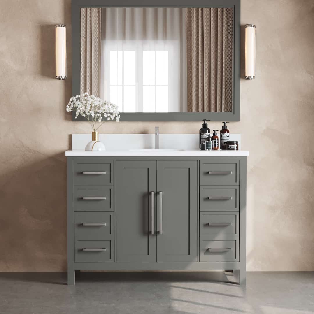 The Acadian 48" Single Bathroom Vanity features a modern design with gray cabinets and a white countertop. A large mirror is complemented by two wall lights mounted on either side, and the spacious counter is adorned with a small arrangement of flowers and various toiletries.