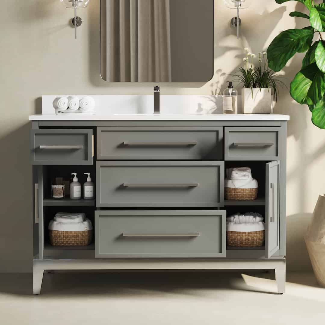 The Barrington 48" Single Sink Bathroom Vanity features a sleek design with gray cabinetry, a spacious countertop, and a large mirror above. Nearby open drawers conveniently display neatly organized towels, baskets, and toiletries against a light wall backdrop, complemented by the presence of a tall green plant.