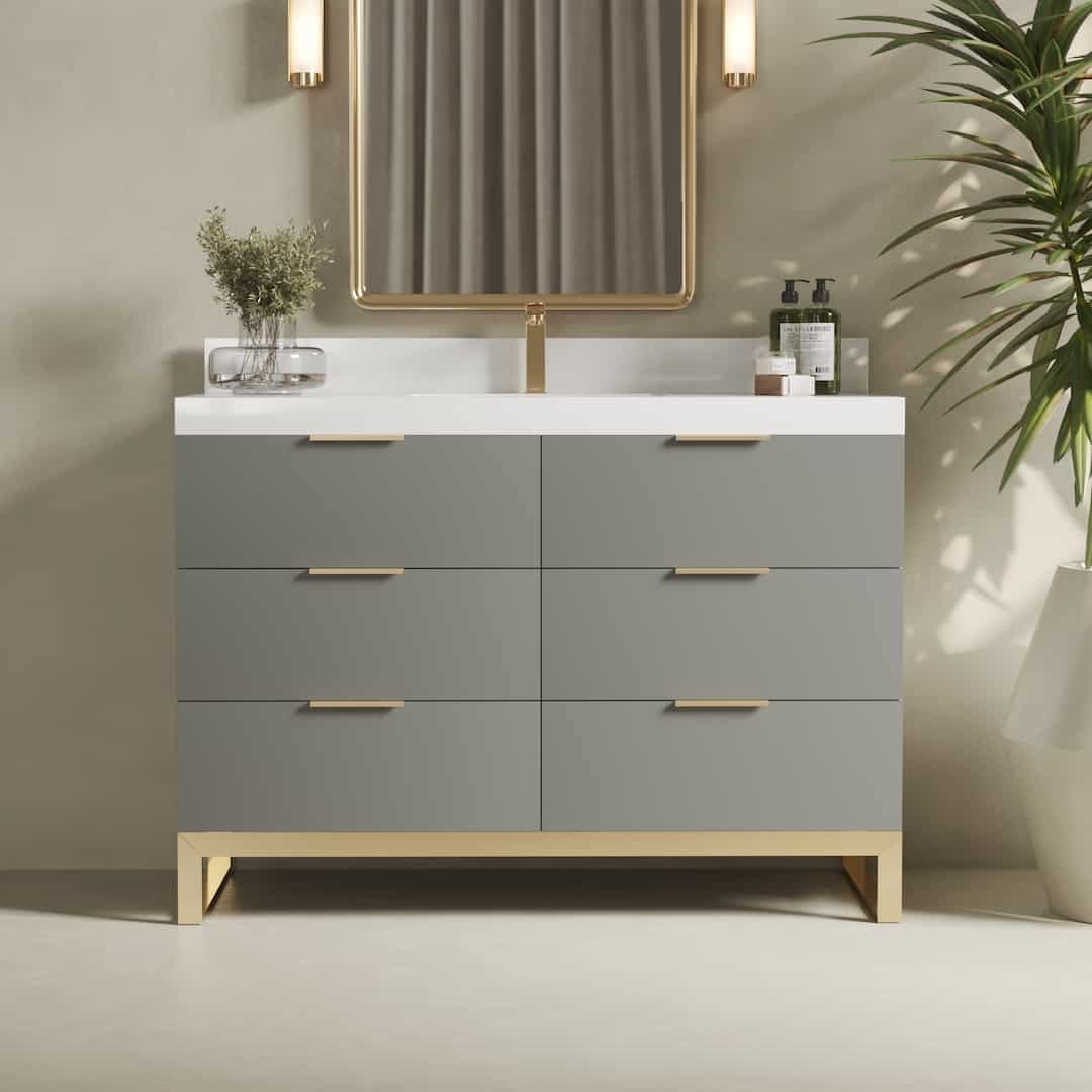 The Haldon 48" Single Sink Bathroom Vanity features a sleek design with six grey drawers adorned with gold hardware, a pristine white countertop, and is complemented by a rectangular mirror. Two stylish wall-mounted lights enhance the modern aesthetic, while a potted plant on the right adds a touch of greenery.