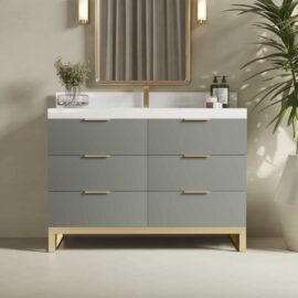 The Haldon 48" Single Sink Bathroom Vanity features a sleek design with six grey drawers adorned with gold hardware, a pristine white countertop, and is complemented by a rectangular mirror. Two stylish wall-mounted lights enhance the modern aesthetic, while a potted plant on the right adds a touch of greenery.